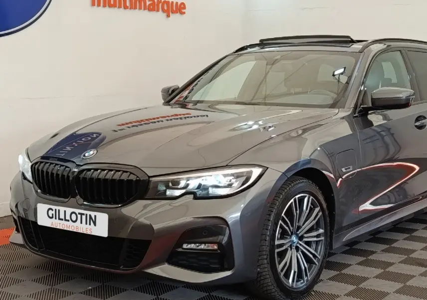 BMW Série 3 320e gris foncé en 3/4 avant droit, avec phares LED allumés et jantes alliage M Sport.