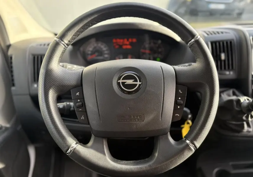 Gros plan sur le volant noir d’un Opel Movano Fourgon 2024 avec commandes intégrées et tableau de bord flou en arrière-plan.