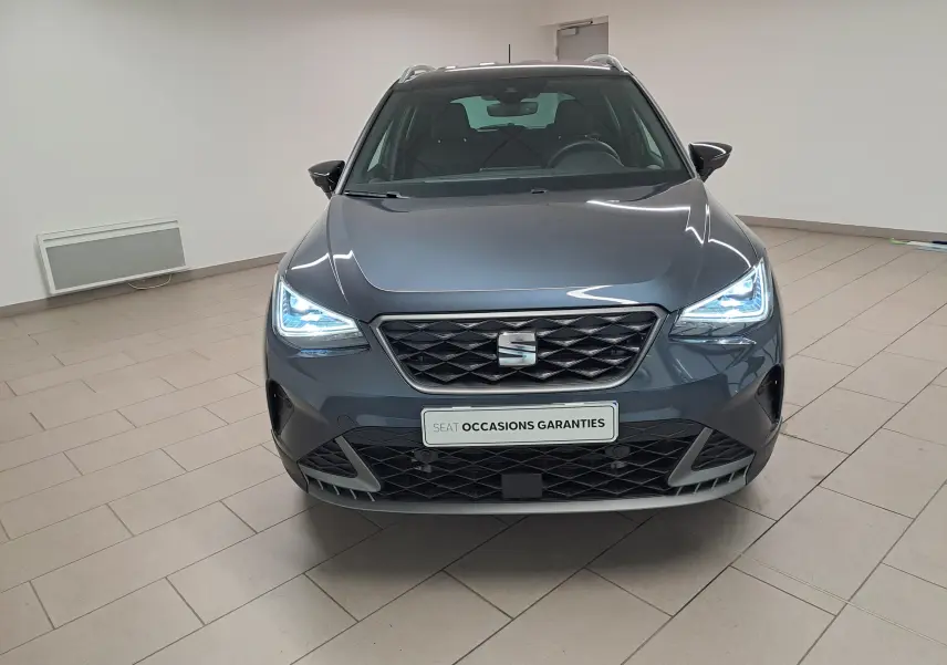 Vue frontale du SEAT Arona 2024 gris magnétique avec toit noir, phares LED allumés dans un showroom.