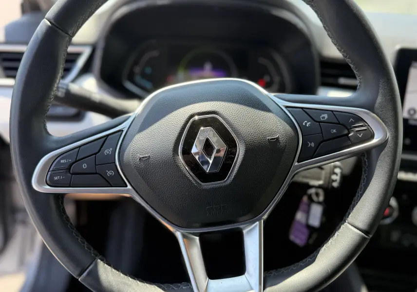 Gros plan sur le volant cuir noir avec logo Renault, tableau de bord numérique flou en arrière-plan dans une Renault Clio V hybride.