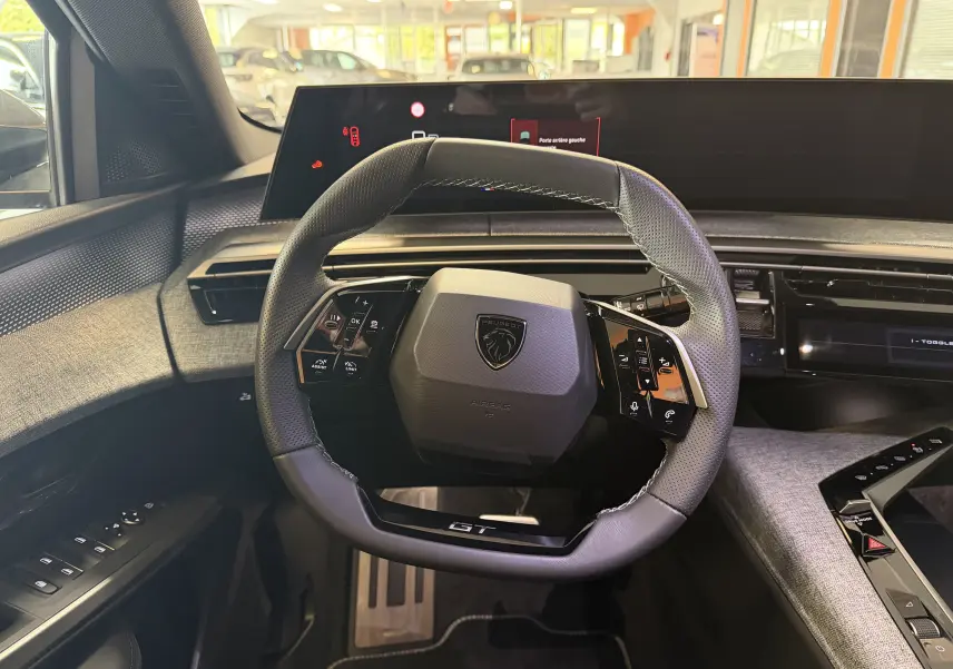 Vue intérieure centrée sur le volant GT du Peugeot 3008 Hybrid 145, avec tableau de bord numérique et habillage tissu gris.