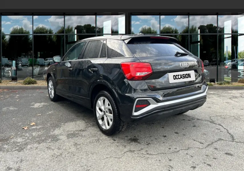 Audi Q2 noir vue 3/4 arrière droit, avec feux arrière rouges et jantes argentées, stationné devant un bâtiment vitré.