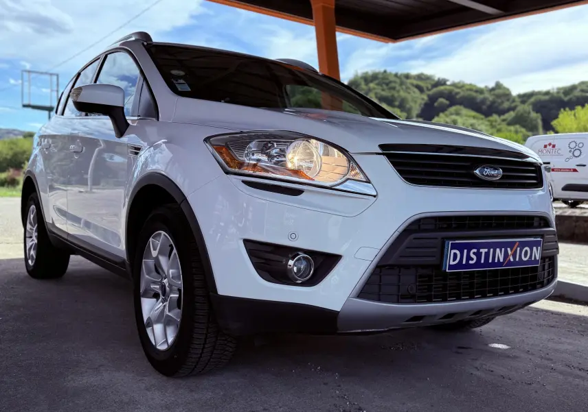 Ford Kuga blanc vu de face droite sous abri, phare avant allumé et calandre noire avec logo Ford.