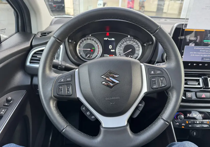 Vue rapprochée du volant noir de la Suzuki S-Cross 1.4 Style Hybrid 2023 avec commandes intégrées et tableau de bord visible.