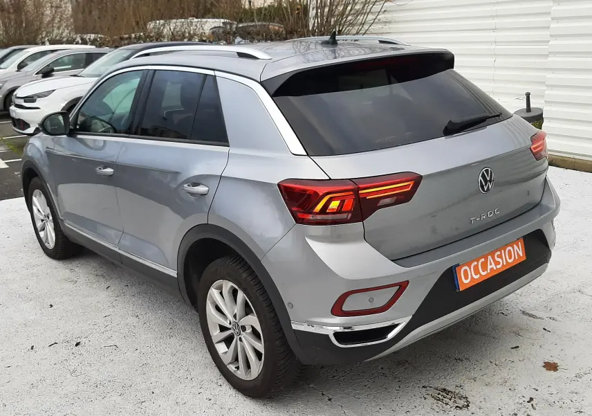 Volkswagen T-Roc gris pyrite vu en 3/4 arrière droit avec feux arrière allumés sur parking extérieur.
