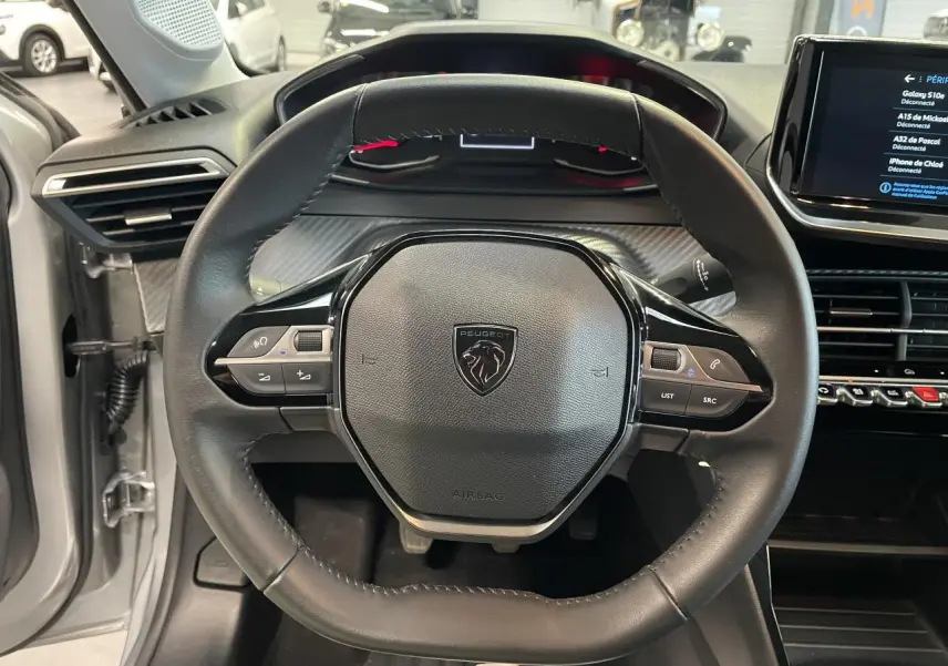 Vue rapprochée du volant cuir noir avec commandes intégrées du Peugeot 208 gris clair, tableau de bord et écran tactile visibles.