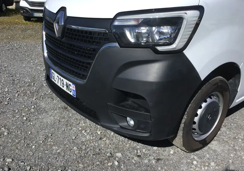 Vue de près 3/4 avant droit du Renault Master blanc avec calandre noire et phare moderne, sur sol gravillonné.