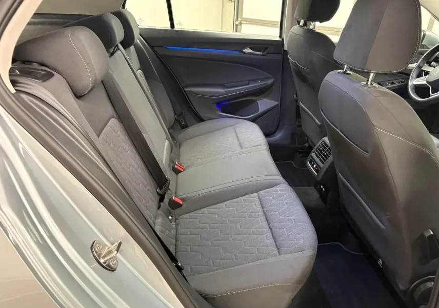 Vue intérieure côté arrière droit de la Volkswagen Golf 2.0 TDI 2025, sièges tissu gris avec éclairage d'ambiance bleu sur la porte.