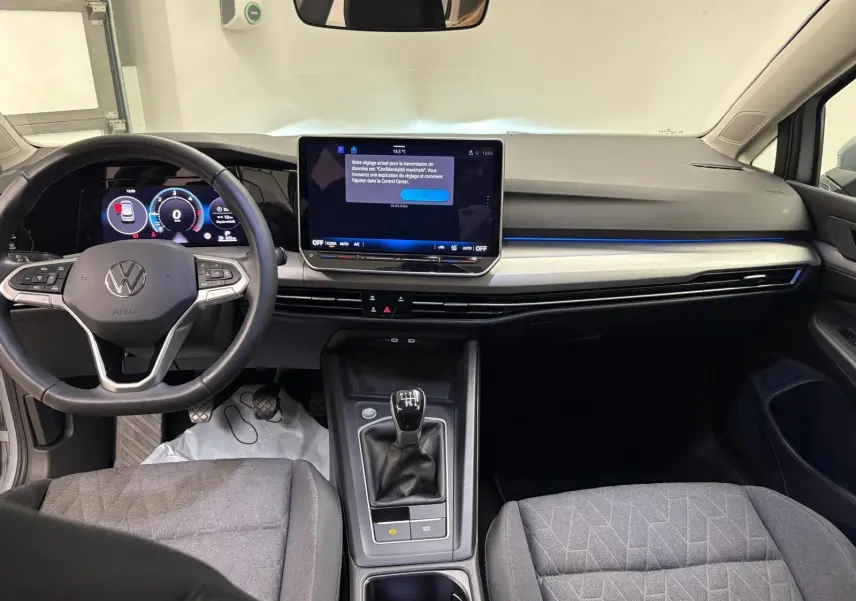 Intérieur de Volkswagen Golf 2025 en vue frontale, tableau de bord noir avec écran tactile et levier de vitesse manuel.