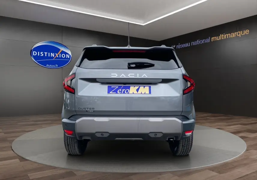 Vue arrière du Dacia Duster hybride gris schiste avec logo distinctif et plaque Zéro KM visible en showroom.