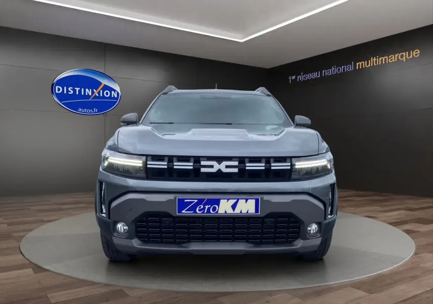 Vue frontale du Dacia Duster gris schiste 2025 avec calandre noire et phares LED allumés en showroom.
