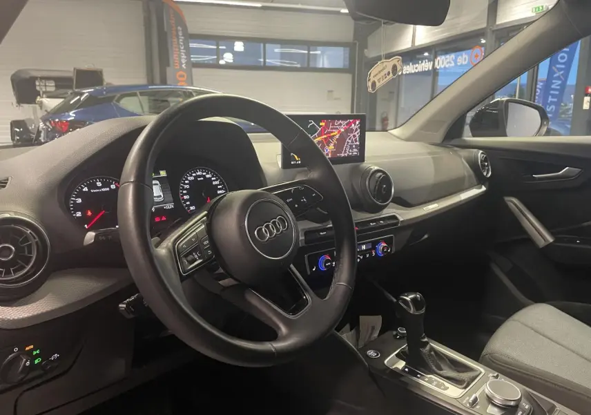 Vue intérieure côté conducteur de l'Audi Q2 2023, volant cuir S line, tableau de bord avec écran navigation et commandes digitales.