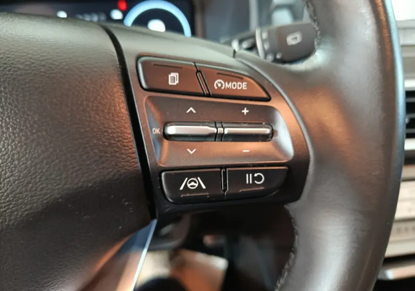 Gros plan sur les commandes du volant cuir noir du Hyundai Kona électrique bleu, avec boutons de régulateur et mode de conduite.