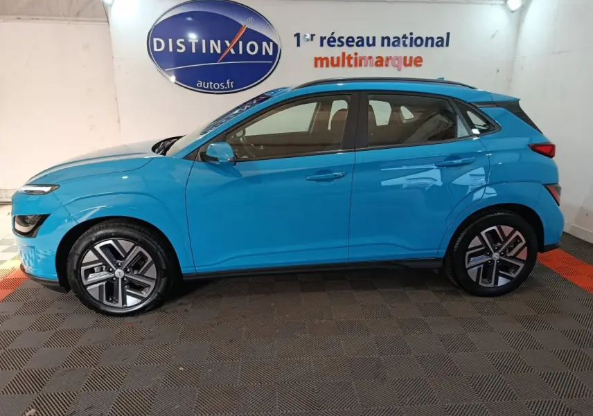 Vue de profil côté gauche d’un Hyundai Kona électrique bleu 2022 stationné en intérieur avec jantes spécifiques et rails de toit noirs.