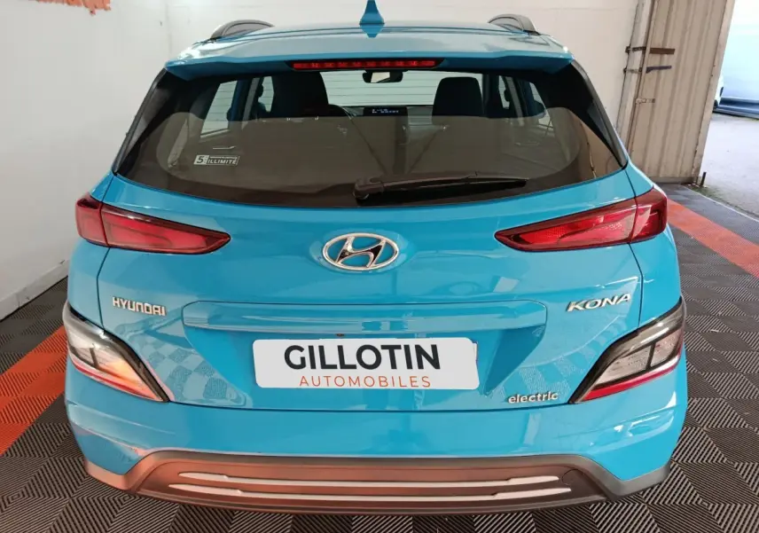 Vue arrière d'une Hyundai Kona électrique bleu vif avec feux arrière allongés et logo visible sur un sol à damier noir et orange.