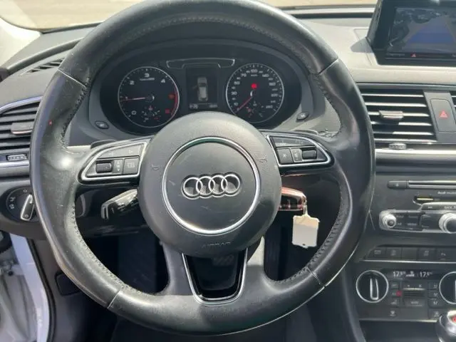 Vue rapprochée du volant en cuir noir d'un Audi Q3 blanc 2015, avec tableau de bord et commandes visibles.