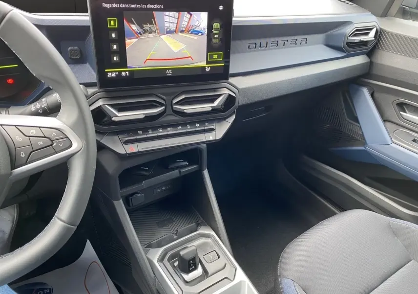 Intérieur du Dacia Duster hybride 2025, tableau de bord noir et bleu avec écran tactile et levier de boîte automatique.