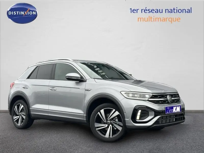 Volkswagen T-Roc gris en 3/4 avant droit avec jantes alu et barres de toit visibles sur fond clair.