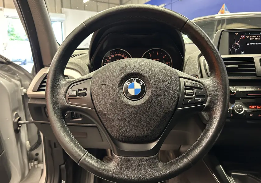 Vue rapprochée du volant BMW Série 1 gris 2014 avec commandes multifonctions et tableau de bord visible.