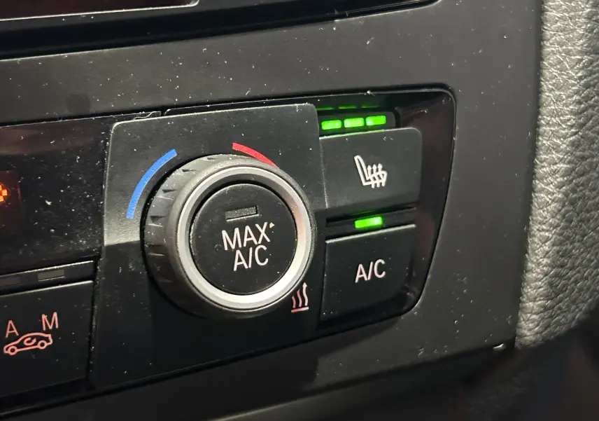 Gros plan sur les commandes de climatisation et siège chauffant activé dans une BMW Série 1 gris diesel de 2014.