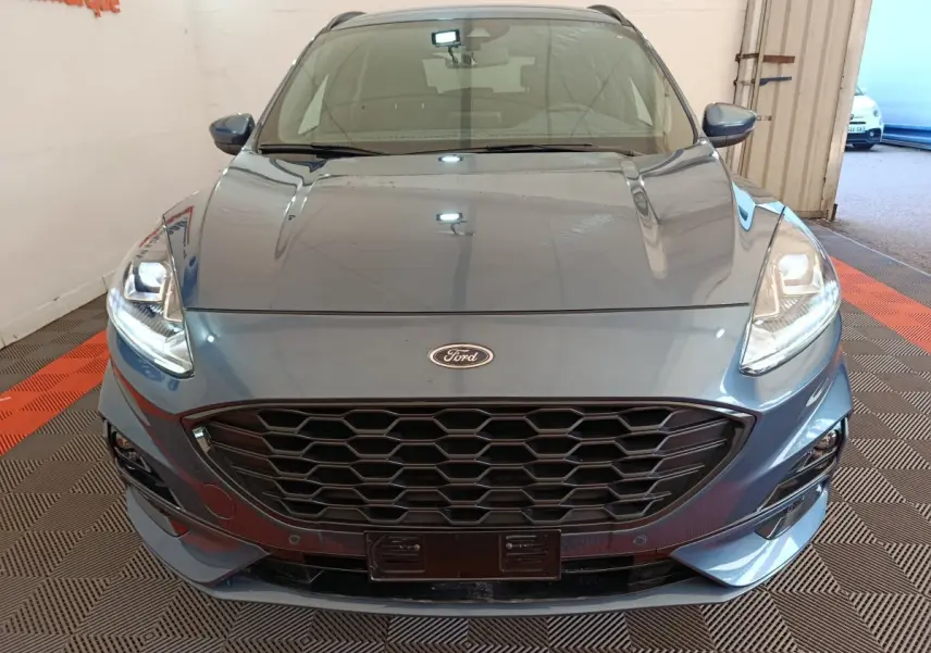 Vue avant rapprochée du Ford Kuga bleu 2024 avec calandre noire et phares allumés dans un intérieur showroom.