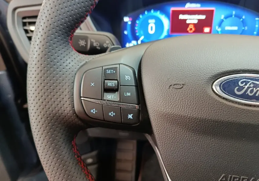 Gros plan sur le volant cuir noir perforé avec commandes multifonctions du Ford Kuga 2024, tableau de bord numérique bleu en arrière-plan.