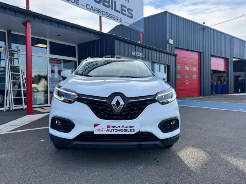 Vue avant d'un Renault Kadjar blanc glacier avec calandre noire et phares LED, stationné devant un garage automobile.