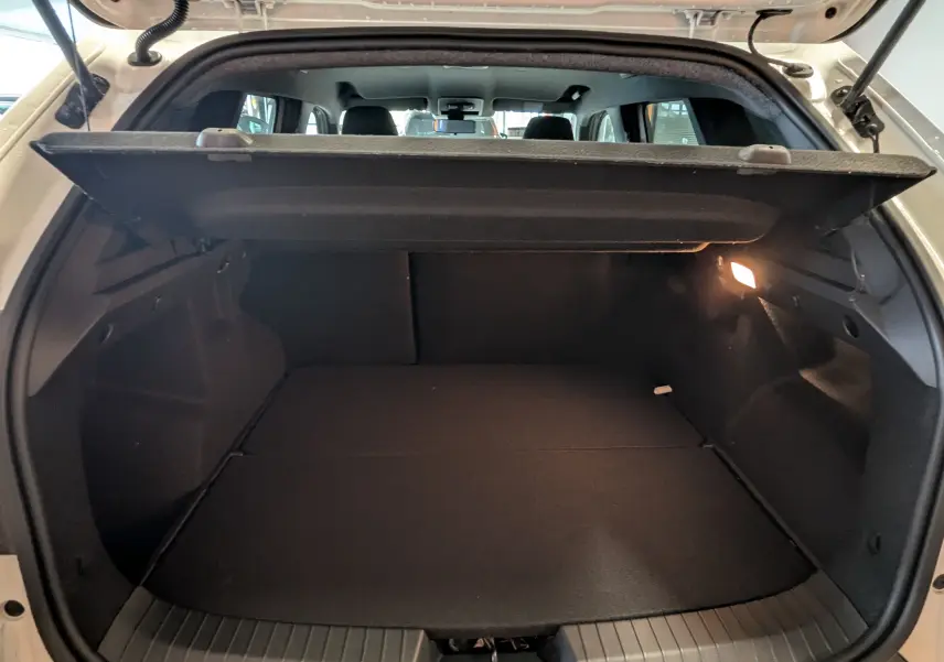 Vue intérieure du coffre ouvert d'un Dacia Duster Hybrid 140 blanc glacier, avec cache-bagages relevé et lumière allumée à droite.