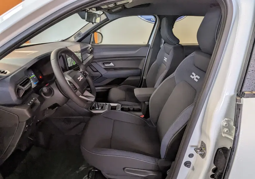 Vue intérieure côté conducteur du Dacia Duster Hybrid 140 Journey blanc glacier, sièges noirs avec logo distinctif sur appuie-tête.