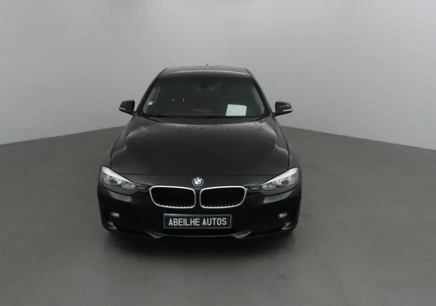 BMW Série 3 316D noire vue de face dans un studio, avec calandre caractéristique et plaque Abeilhe Autos visible.