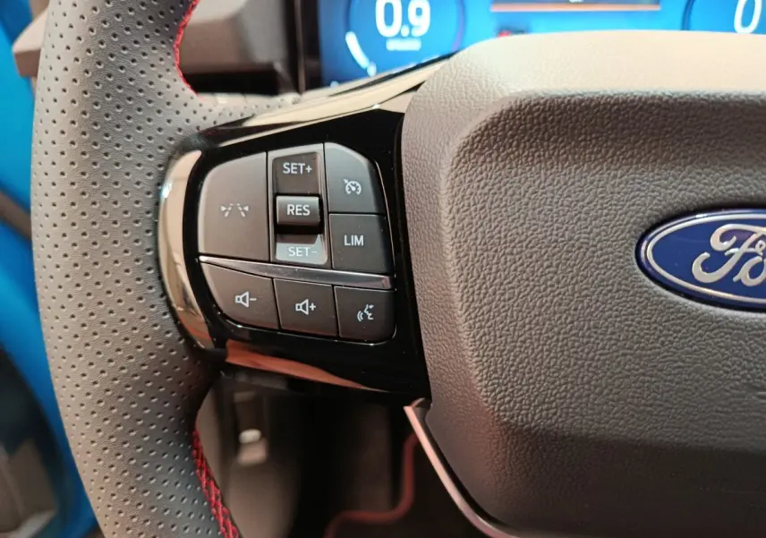 Gros plan sur le volant cuir noir perforé avec commandes multifonctions à gauche du Ford Puma bleu.