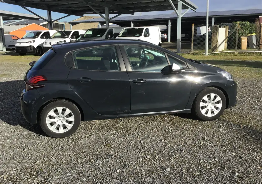 Peugeot 208 noire vue de profil côté gauche, garée sur un sol gravillonné avec des véhicules utilitaires en arrière-plan.