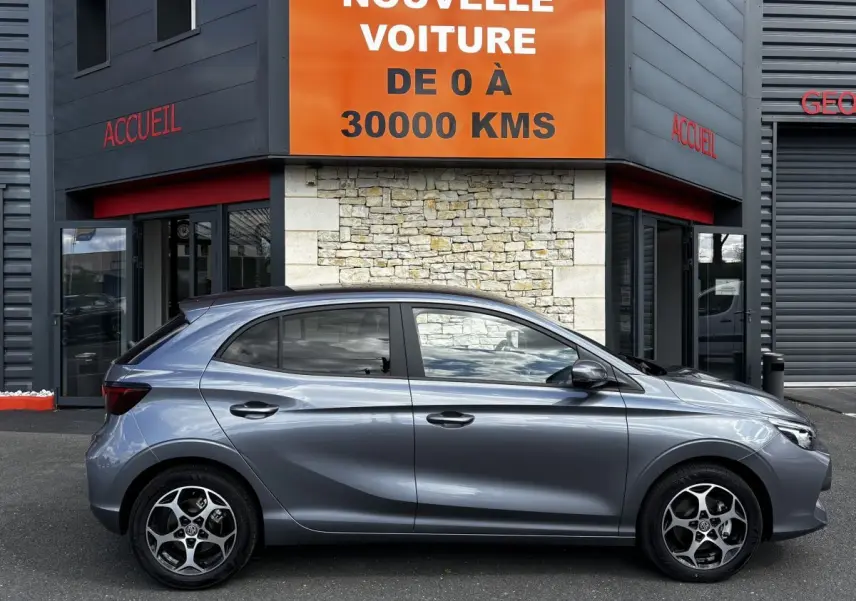 Vue de profil côté gauche d'une MG3 gris clair 2025 garée devant un bâtiment avec un panneau orange "Votre nouvelle voiture".