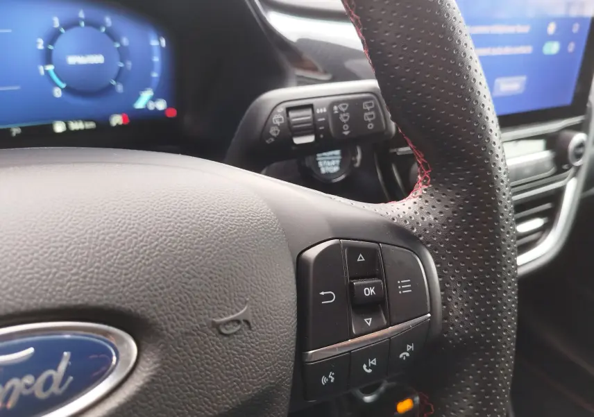 Gros plan sur le volant en cuir perforé avec surpiqûres rouges du Ford Puma 1.0 EcoBoost, tableau de bord numérique en arrière-plan.