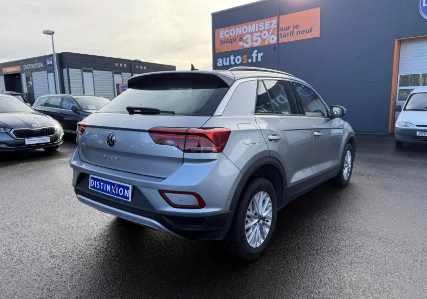 Volkswagen T-Roc gris pyrite vu en 3/4 arrière droit sur parking devant un bâtiment commercial.