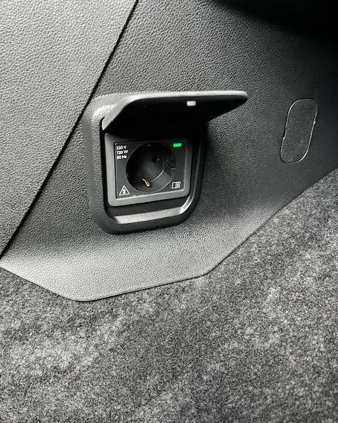 Prise 220V intégrée dans le coffre du Peugeot Rifter gris foncé, avec trappe de protection ouverte.