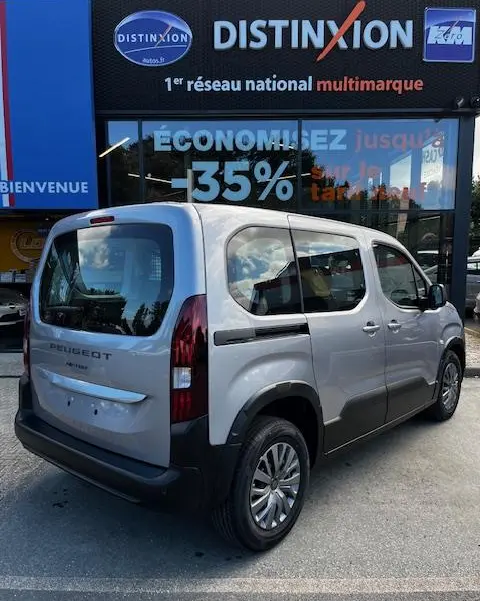 Vue 3/4 arrière droite d’un Peugeot Rifter gris foncé 2024 avec vitres opaques et rails de toit, stationné devant une concession multimarque.