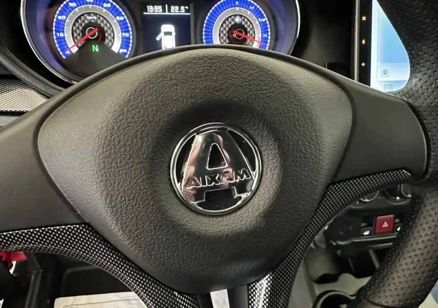 Vue rapprochée du volant noir avec logo Aixam, tableau de bord bleu et écran tactile partiel à droite.