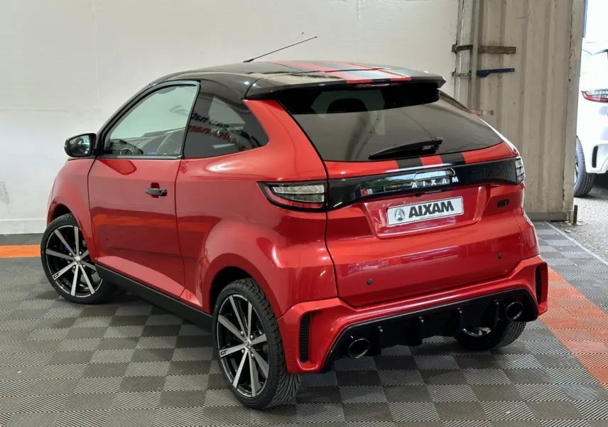 Vue 3/4 arrière droite d'une Aixam Ambition rouge 2026 avec toit noir, jantes noires et bandes rouges sur le toit.