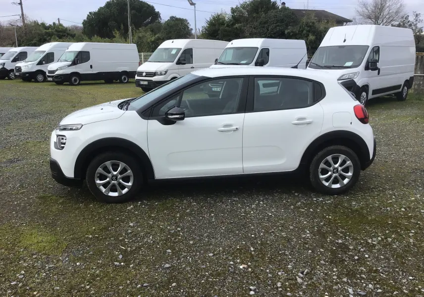 Citroën C3 blanc vue de profil côté gauche, avec jantes alliage et entourée de fourgons blancs en arrière-plan.