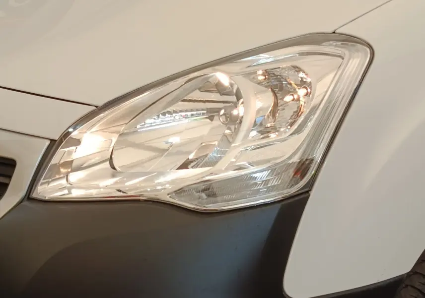 Gros plan sur le phare avant droit du Peugeot Partner blanc, avec partie inférieure du pare-chocs noir visible.