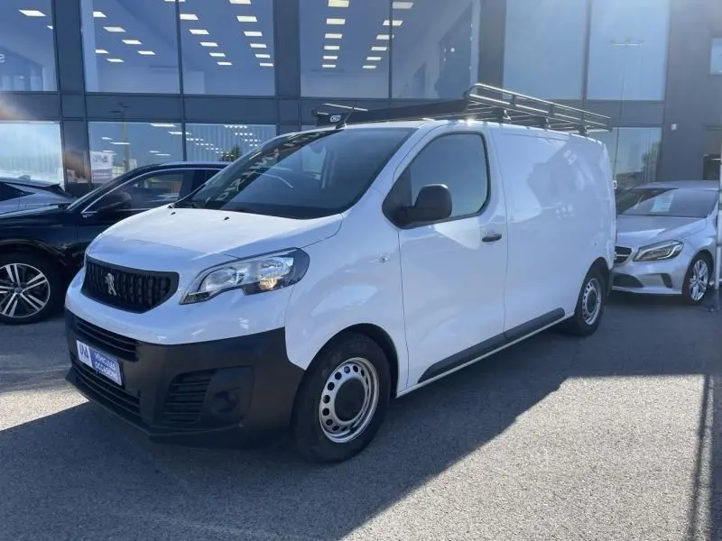 Peugeot Expert Fourgon blanc en 3/4 avant droit, avec calandre noire et jantes acier simples, stationné devant un bâtiment vitré.