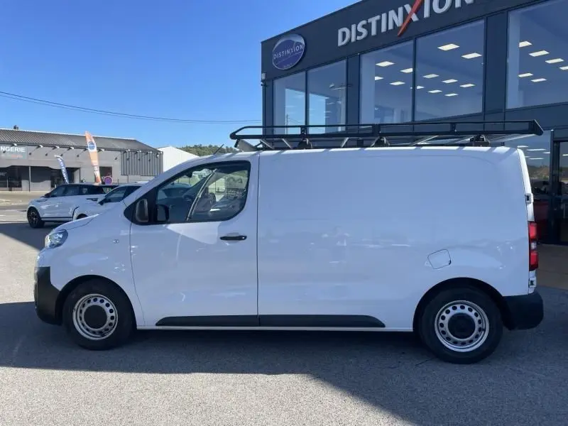 Vue latérale côté droit d'un Peugeot Expert Fourgon blanc 2023 avec galerie de toit noir, stationné devant un bâtiment commercial.
