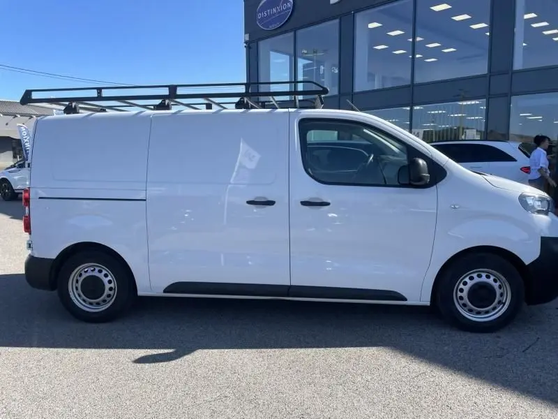 Vue de profil côté droit d'un Peugeot Expert Fourgon blanc 2023 avec galerie de toit noire sur un parking.