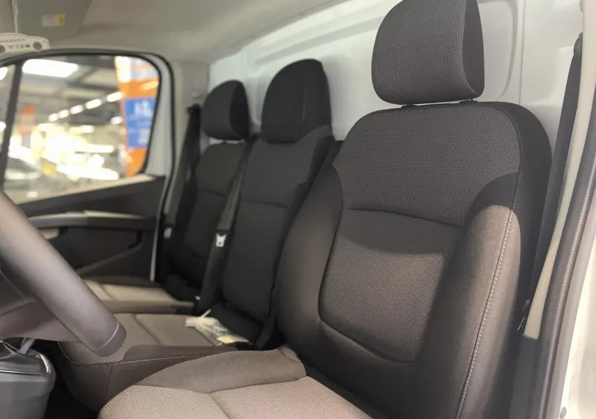 Vue intérieure côté conducteur du Renault Trafic Fourgon blanc, montrant les sièges noirs en tissu et la banquette passager.