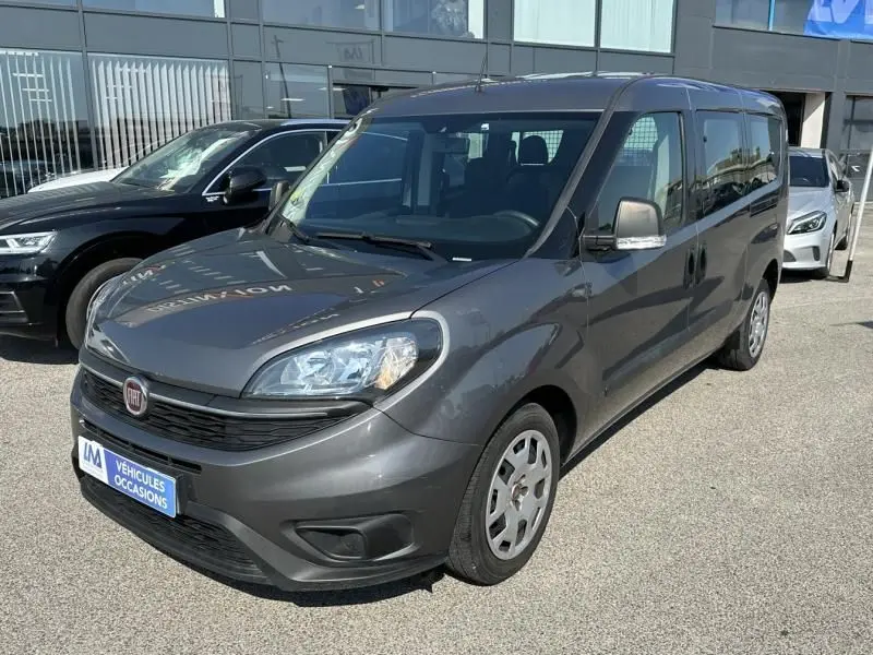 Fiat Doblo Cargo gris foncé vu en 3/4 avant droit sur parking devant un bâtiment vitré.