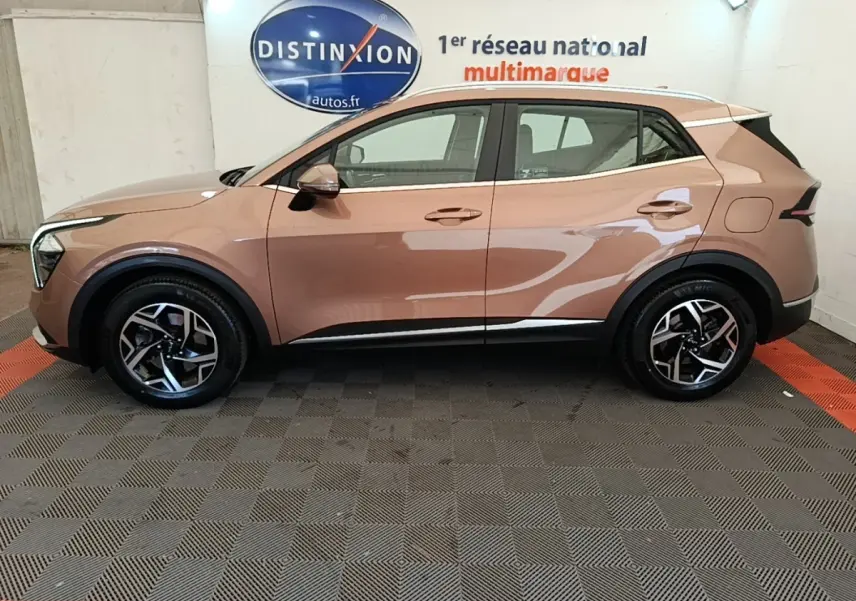 Vue latérale droite d'un KIA SPORTAGE beige 2022 avec jantes alliage et rails de toit noirs en intérieur showroom.