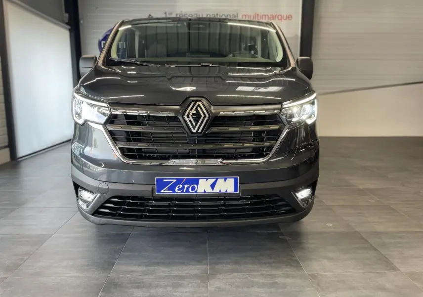 Vue frontale d'un Renault Trafic Fourgon gris Comète avec phares et feux de jour allumés en intérieur showroom.