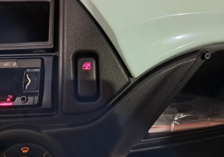 Gros plan sur le bouton de commande de lève-vitre avec tableau de bord noir et partie de la carrosserie verte côté conducteur.