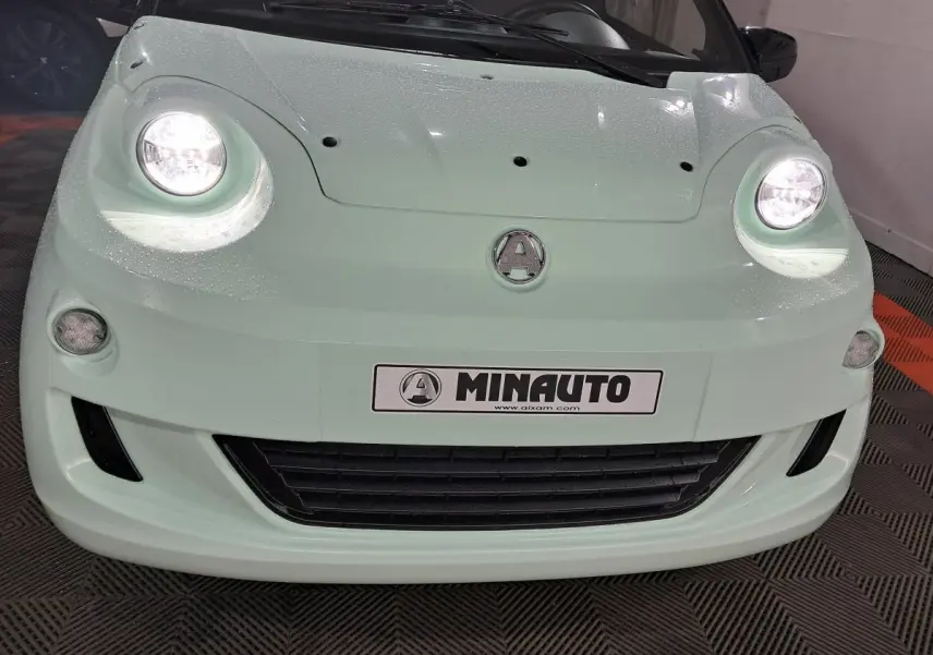 Vue avant rapprochée de l'Aixam Minauto E chic 2026 vert clair avec phares allumés et logo central visible.